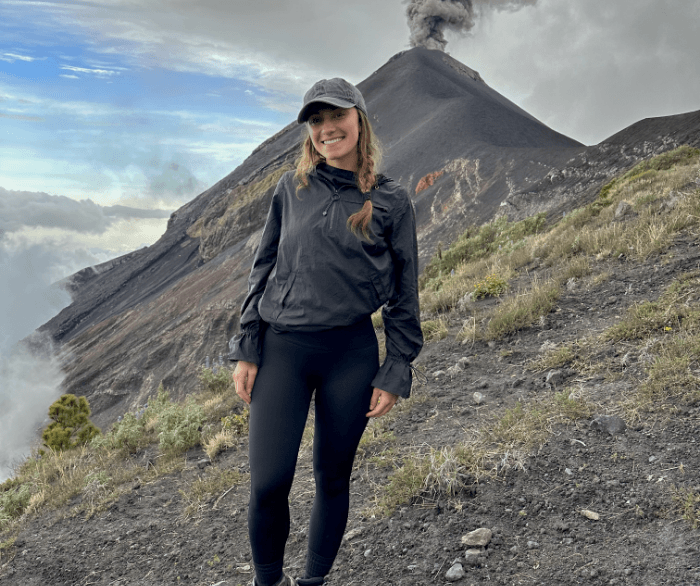 Dr. Antonella Barrios stands on a mountain, wearing a black jacket and pants, with a volcano visible in the background